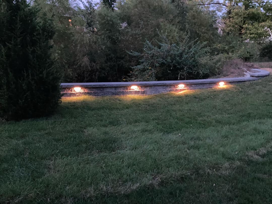 Outdoor landscape with illuminated stone wall and greenery at dusk.