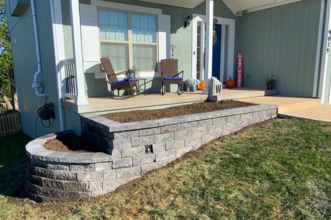 Stone retaining wall landscaping in front of a house with chairs and fall decorations.