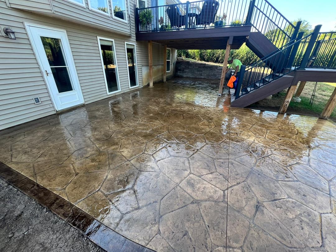Stamped concrete patio with a worker cleaning the surface beside a two-story house.