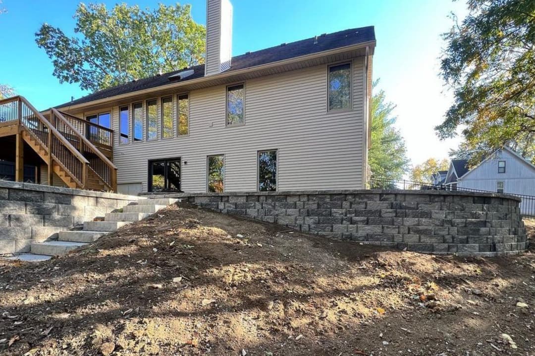 Modern home exterior with large windows, stone wall, and wooden deck in a wooded setting.