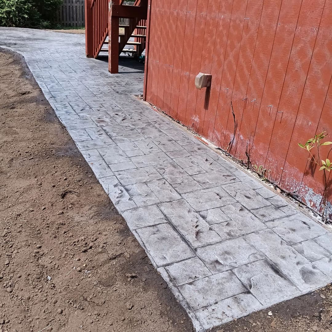 Stamped concrete walkway leading to a red house, with bare soil on the sides.