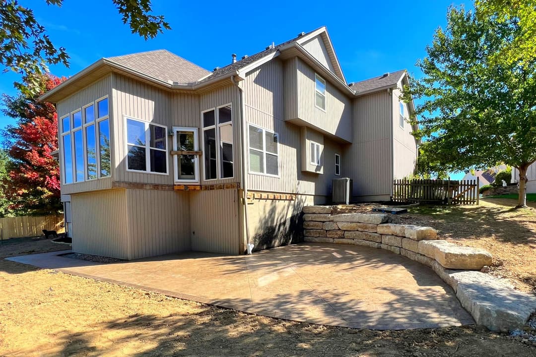 Modern home with large windows, stone patio, and landscaped yard on a sunny day.