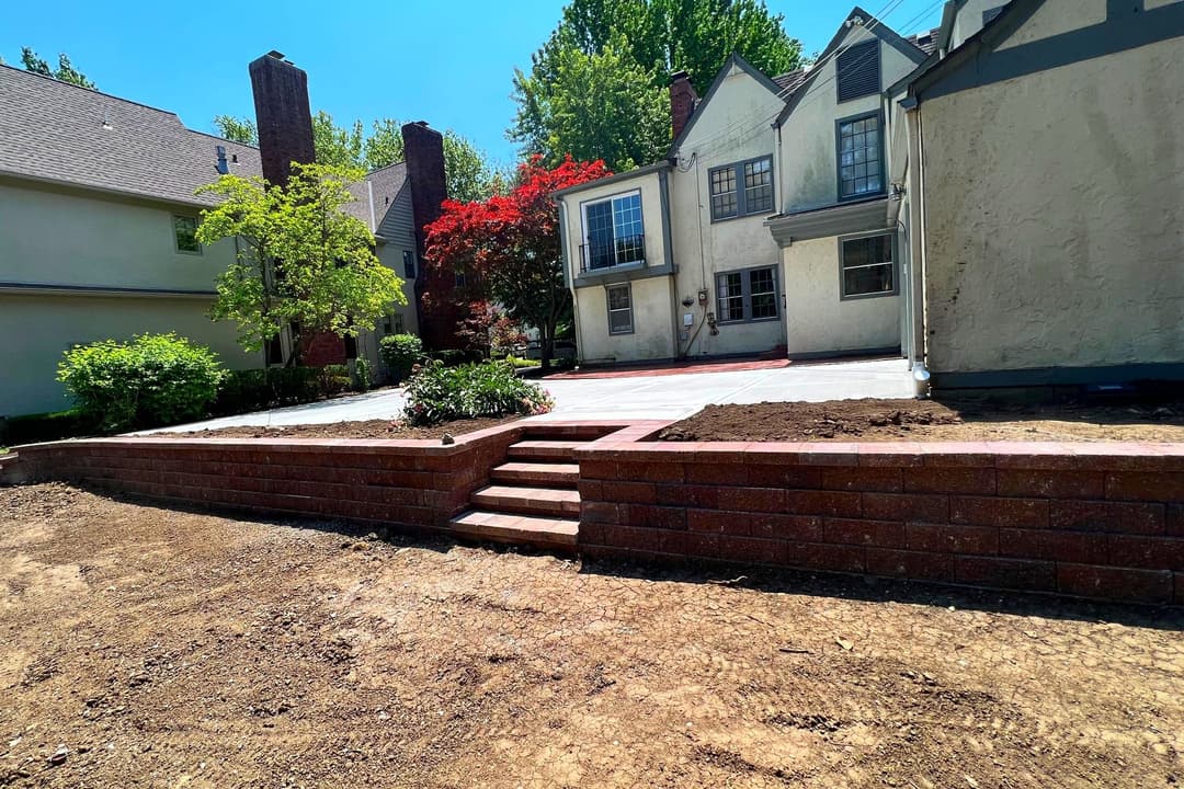 Residential backyard with brick patio and steps, surrounded by green landscaping.